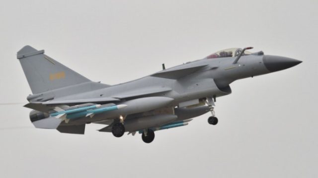 Jet tempur Chengdu J-10CE berwarna abu-abu muda sedang lepas landas atau mendarat dengan roda pendarat terbuka dan beberapa rudal terpasang di bawah sayapnya, menunjukkan kesiapan tempur